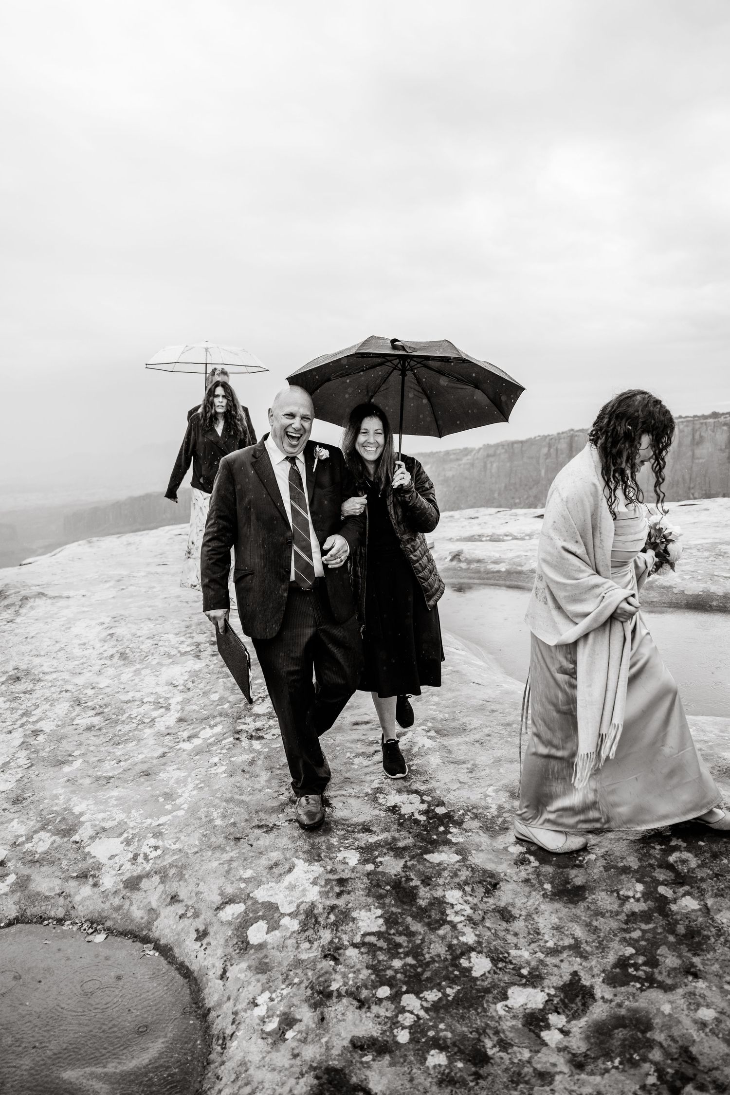 Wedding group navigating wet rocky terrain with umbrellas in black and white photography.