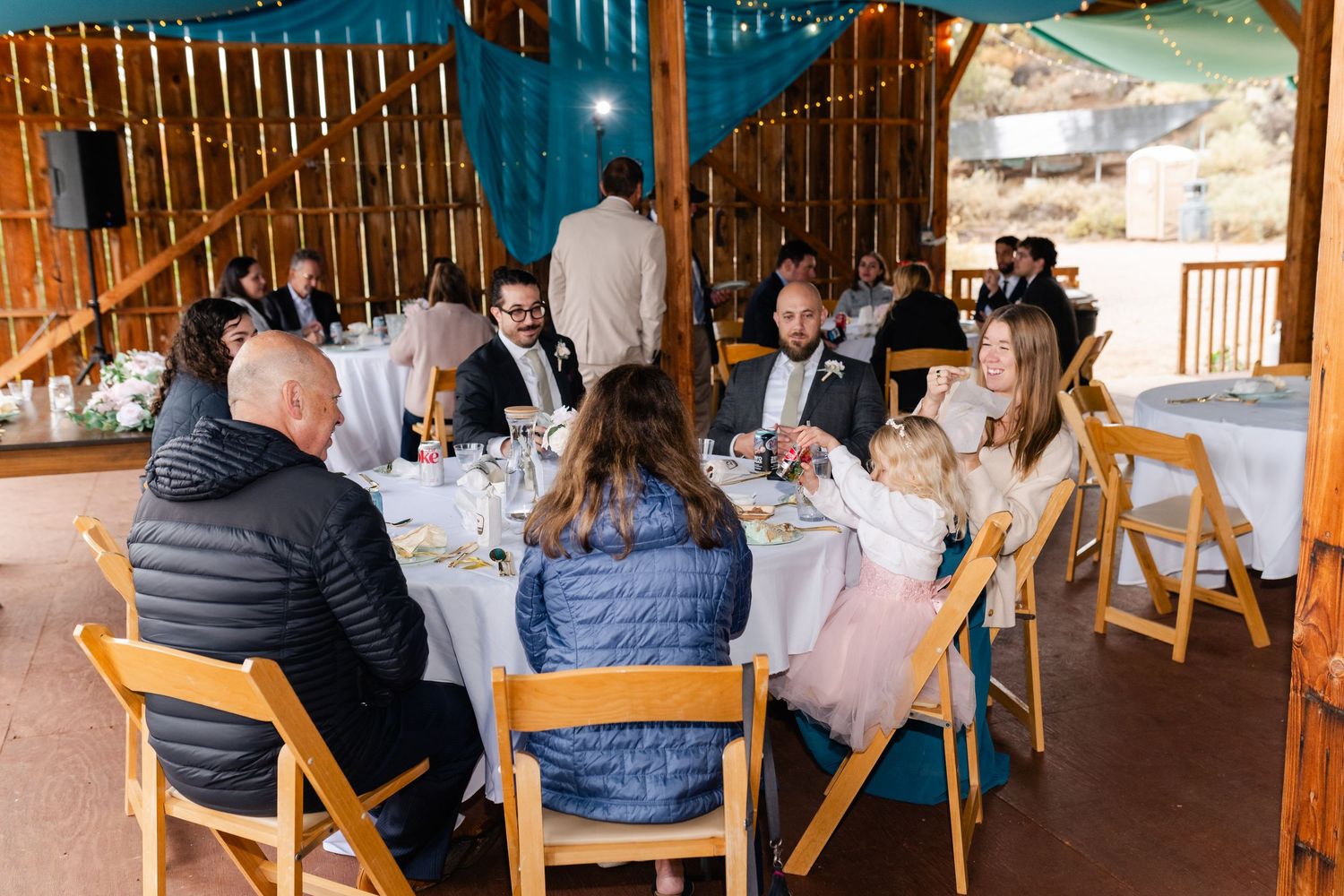 Wedding guests gathered around tables for a meal under a covered outdoor venue.