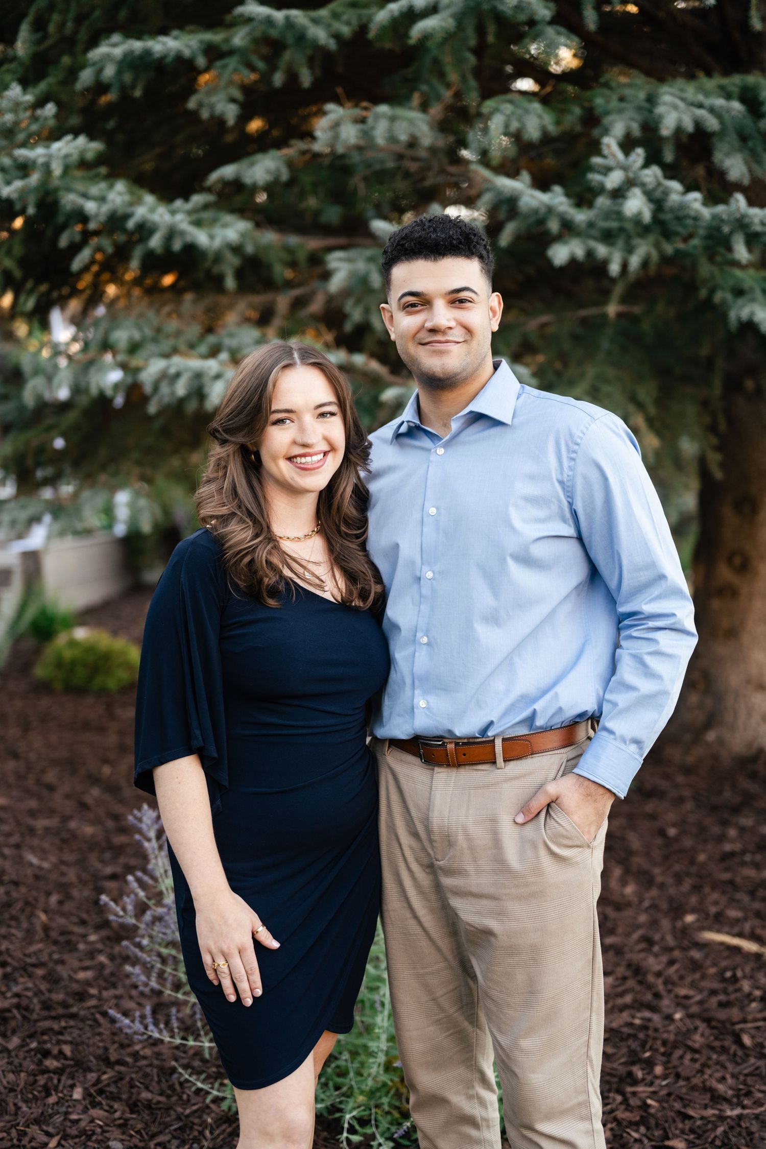 Couple in coordinating navy and light blue outfits pose together for casual outdoor photos among evergreen trees.
