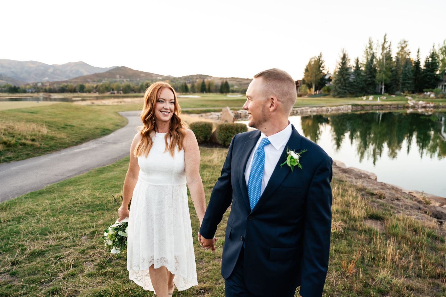 A couple enjoying their wedding day by a serene lake with mountains and evergreen trees in the background.