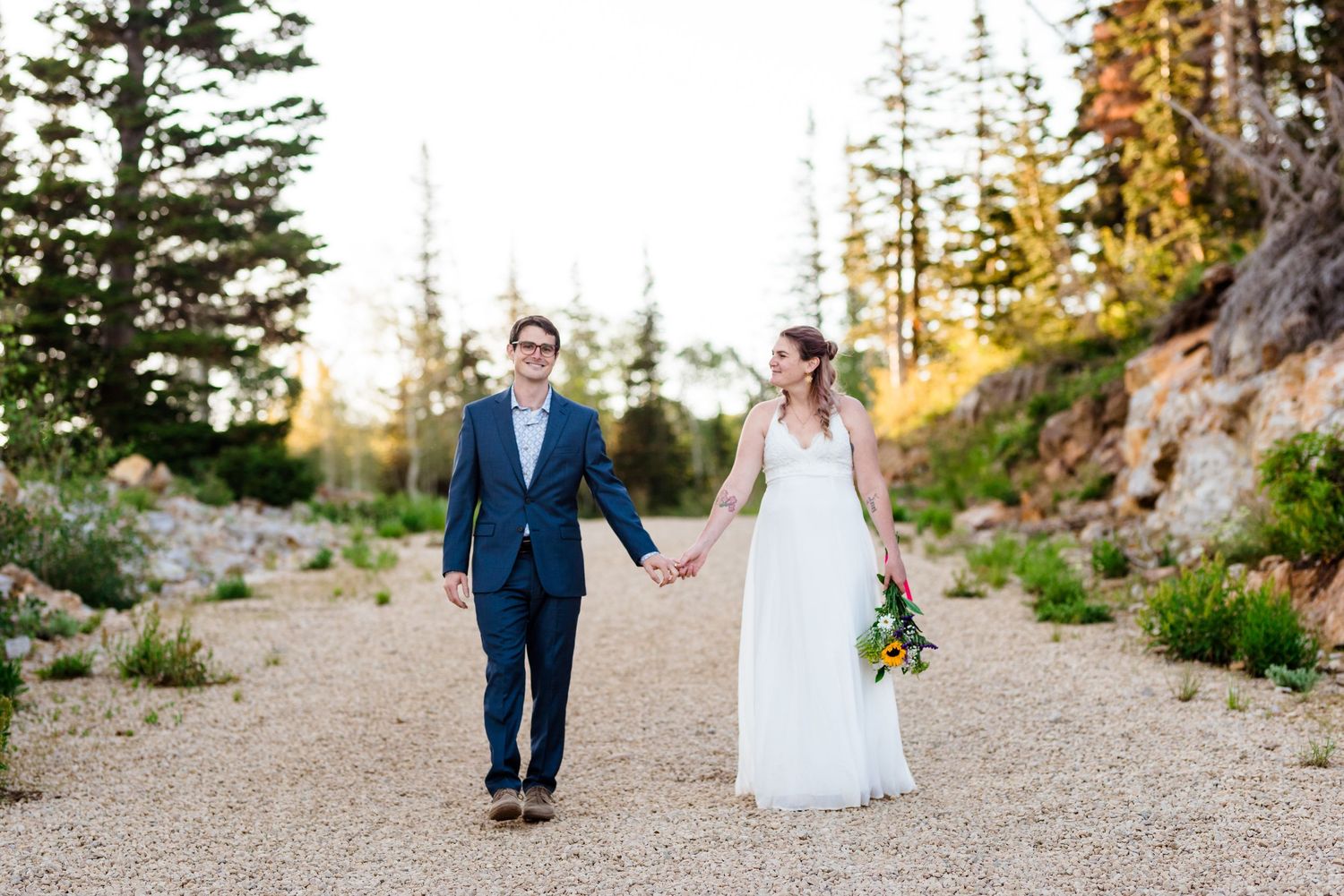 A couple walks hand in hand on a mountain path at sunset, surrounded by evergreen trees and wildflowers.