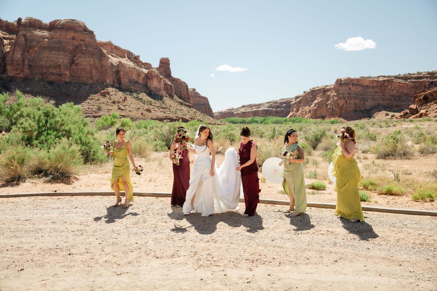 Bride and bridesmaids in burgundy and yellow dresses posing in a desert canyon landscape on a sunny wedding day.