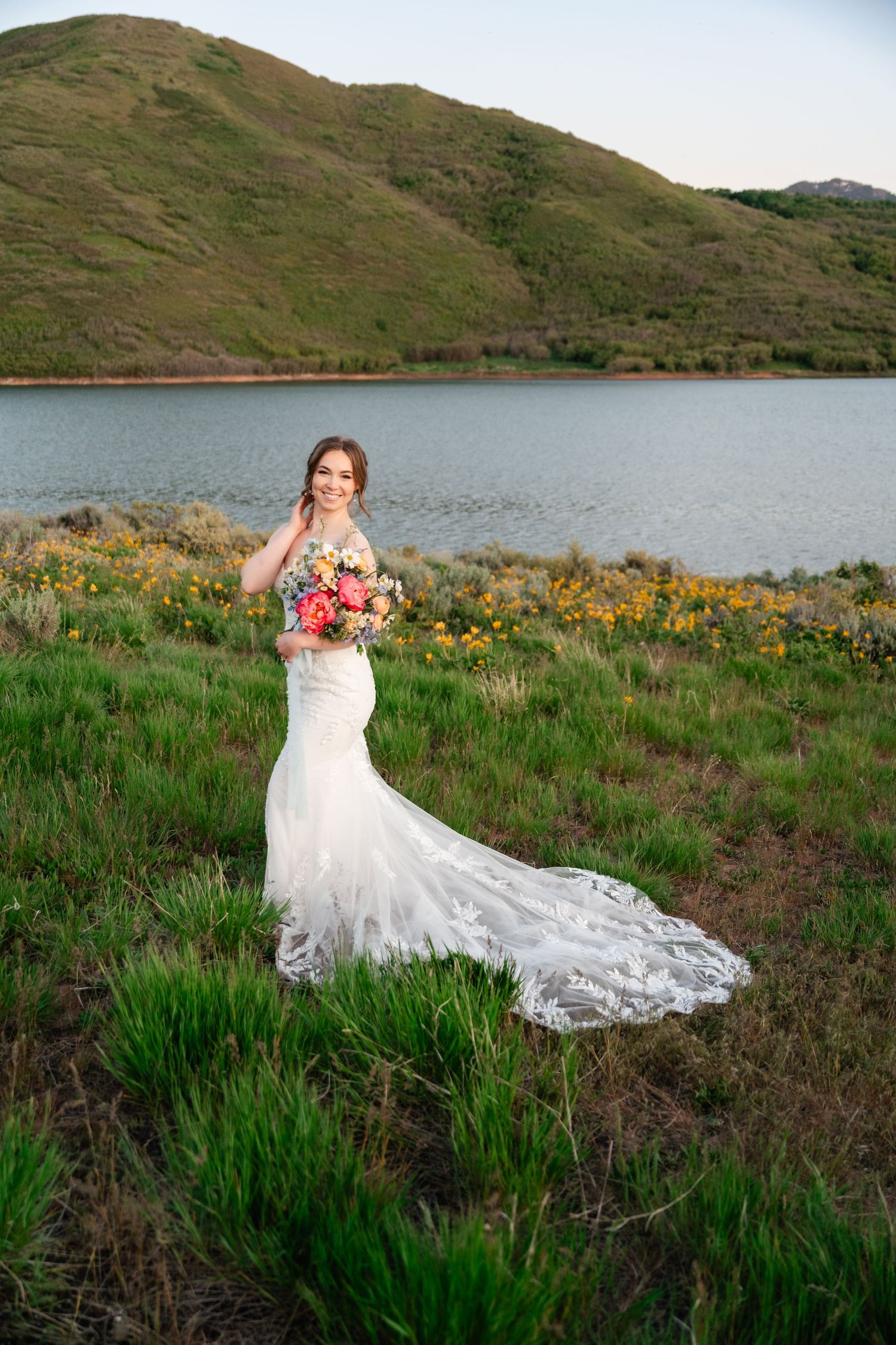 White wedding dress with lace train flows across green meadow near mountain lake at sunset.