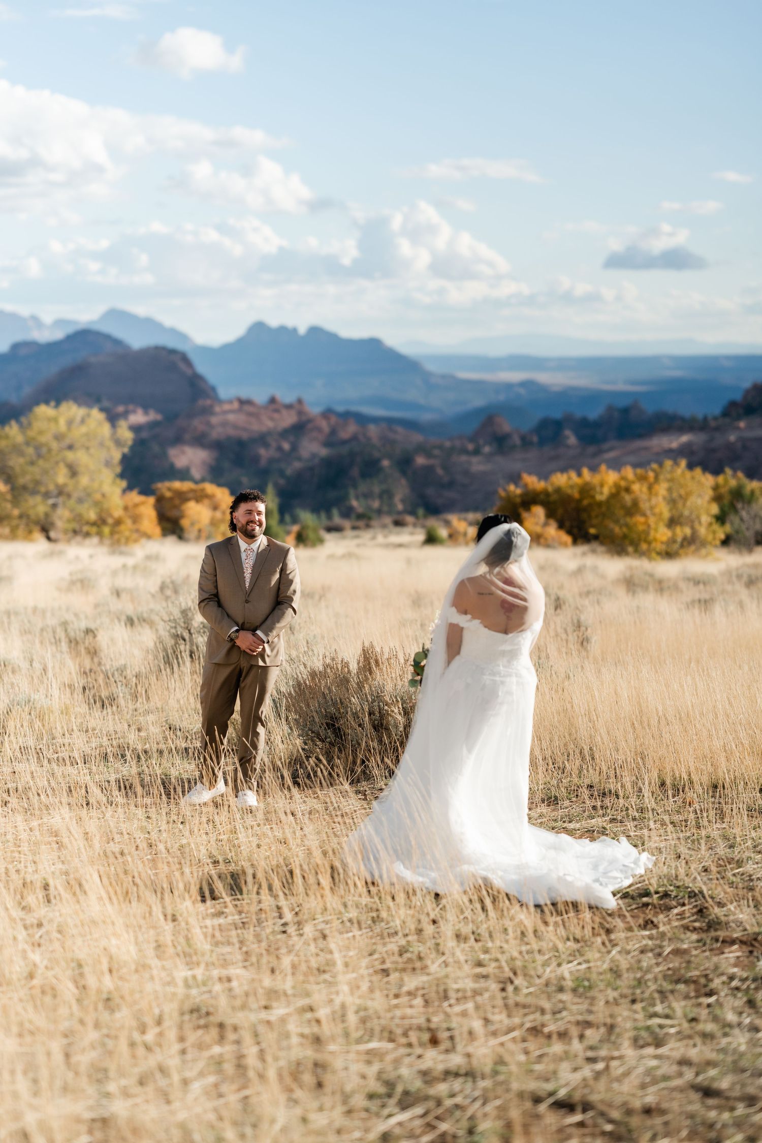 Wedding couple poses playfully in a golden field with dramatic mountain backdrop at sunset.