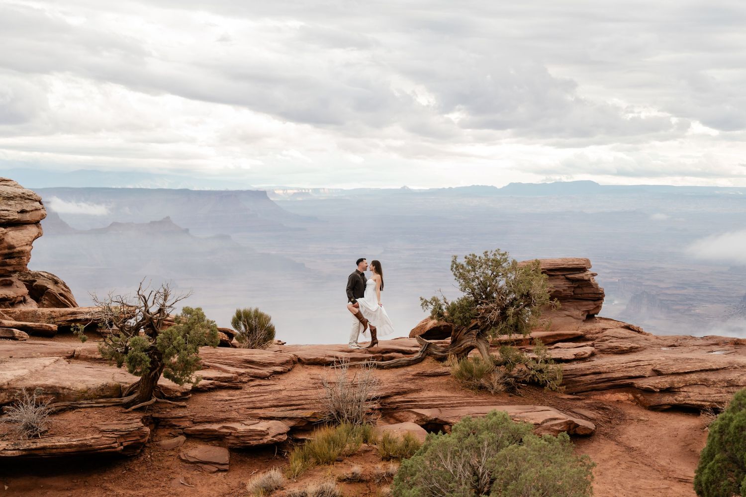 A couple stands on dramatic red rock cliffs overlooking a vast canyon vista.