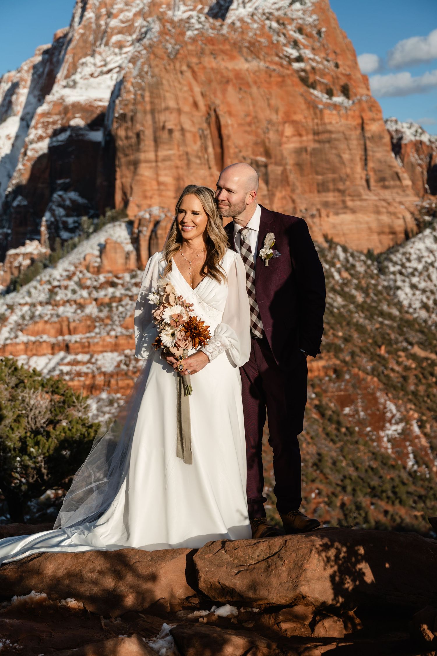 A wedding couple poses in front of majestic red rock cliffs during golden hour.