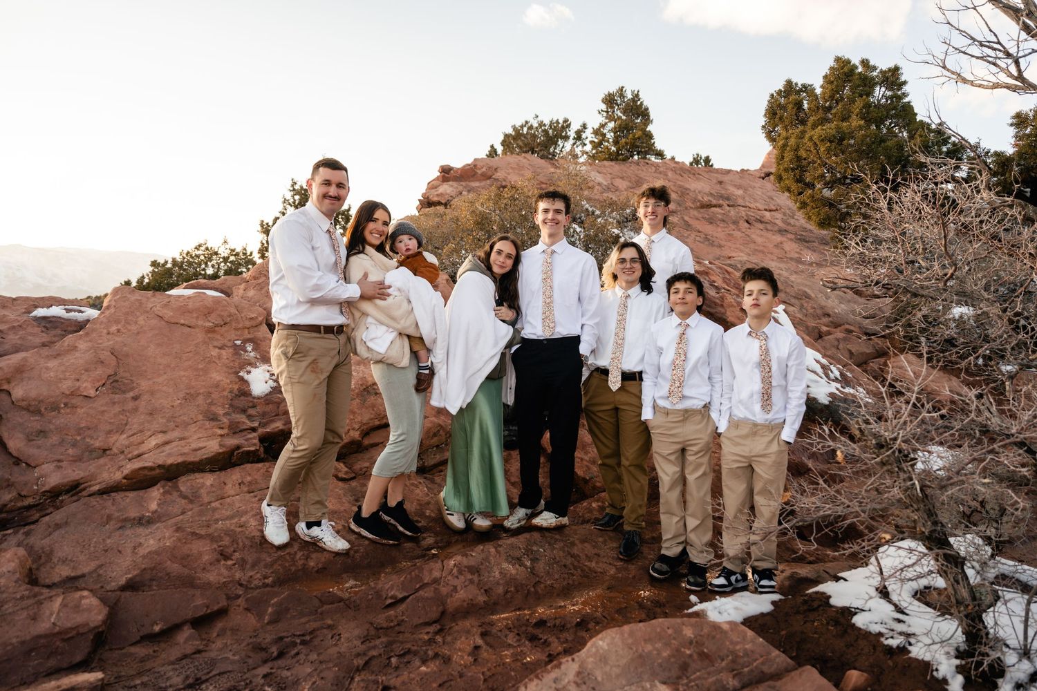Wedding party in white and neutral attire poses along red rock cliff edge with mountain backdrop.