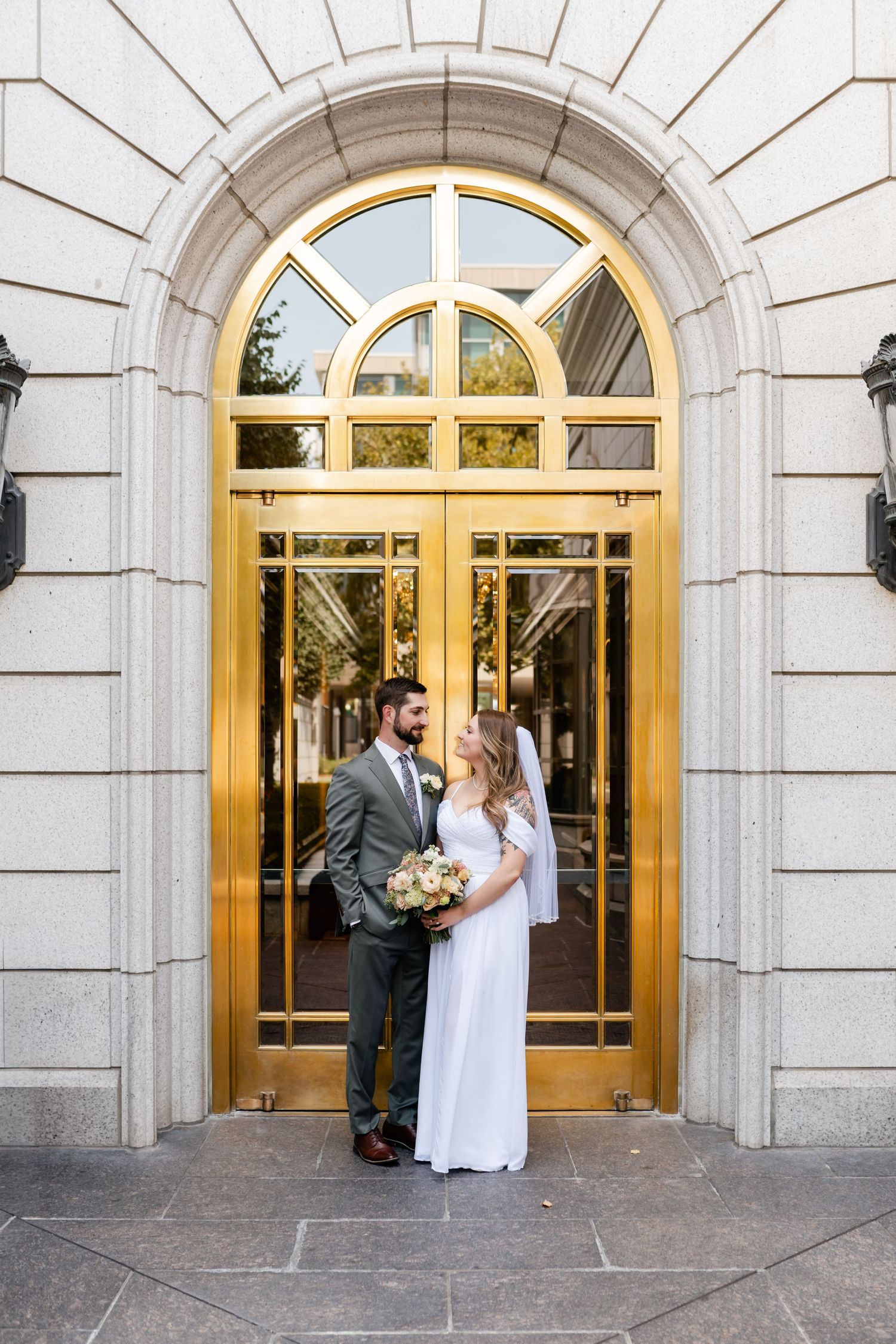 A couple stands in front of grand arched golden doors at an elegant stone building entrance.