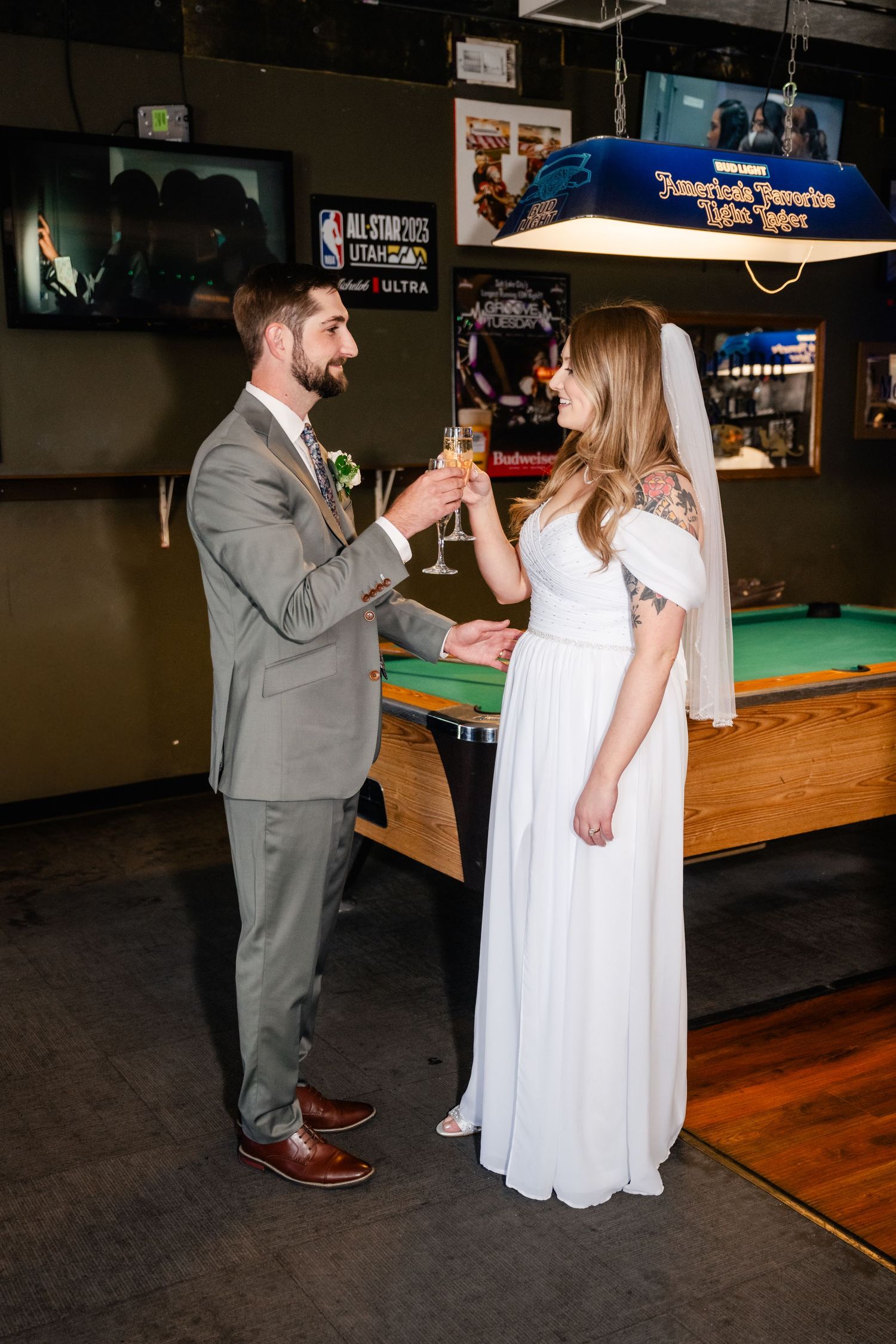 A couple in wedding attire sharing a toast next to a pool table in a casual bar setting.