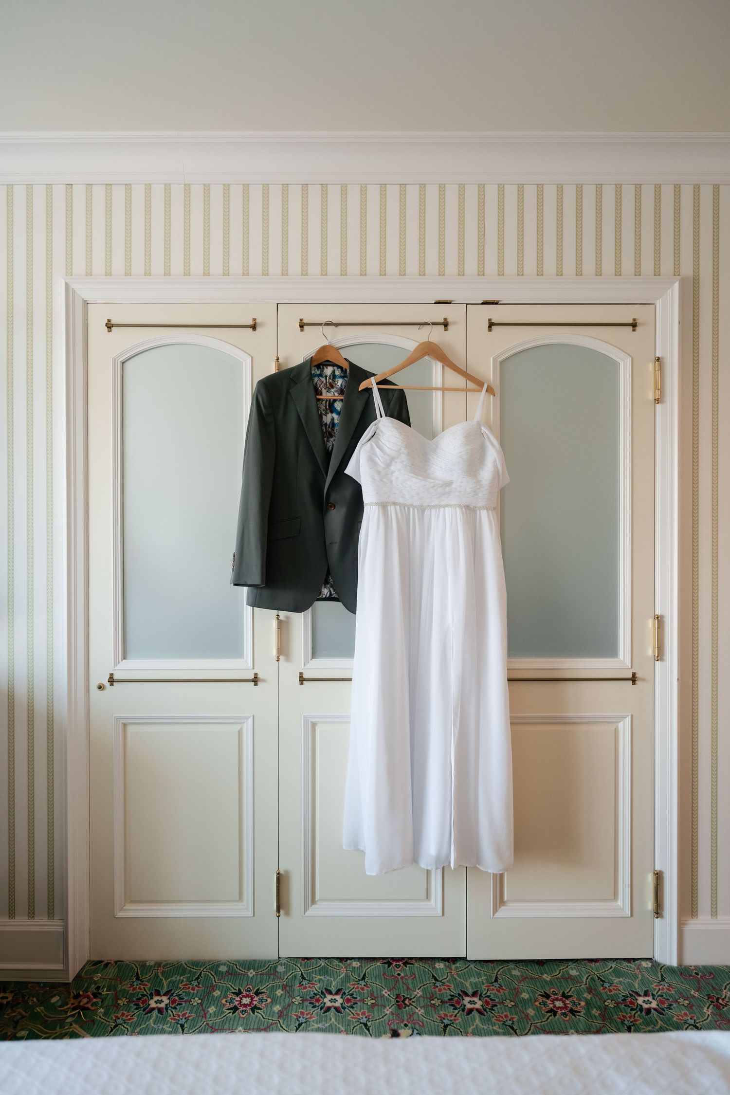 Wedding attire including a black suit and white dress hanging on wooden hangers against a cream colored wall.
