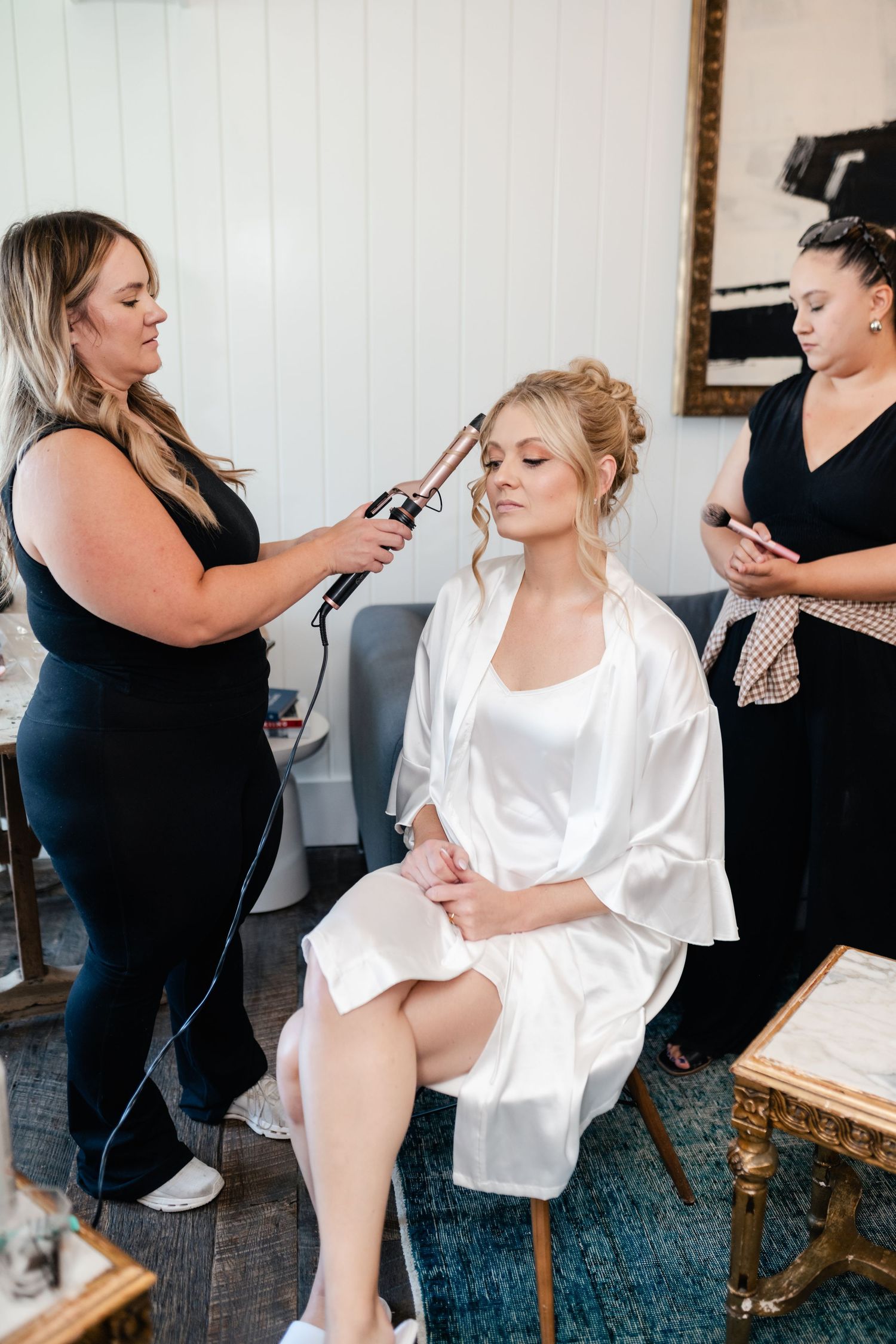 Hairstylist applies makeup to client in white silk robe while another stylist checks phone during wedding preparation.