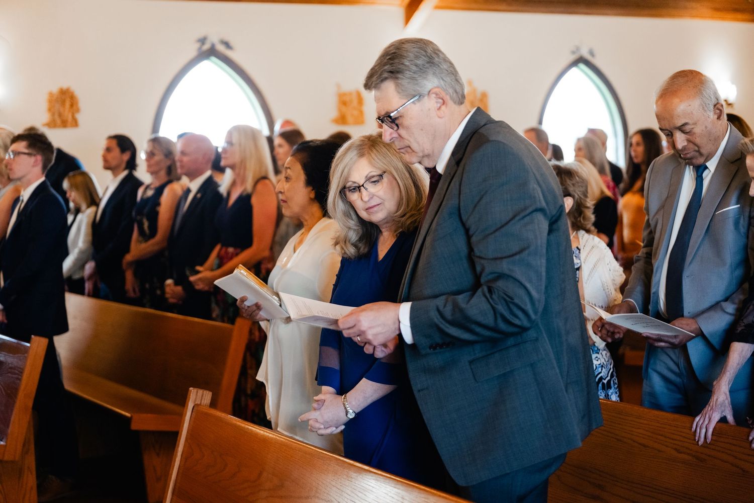Wedding guests sit in wooden church pews while reading from programs during a traditional ceremony service.