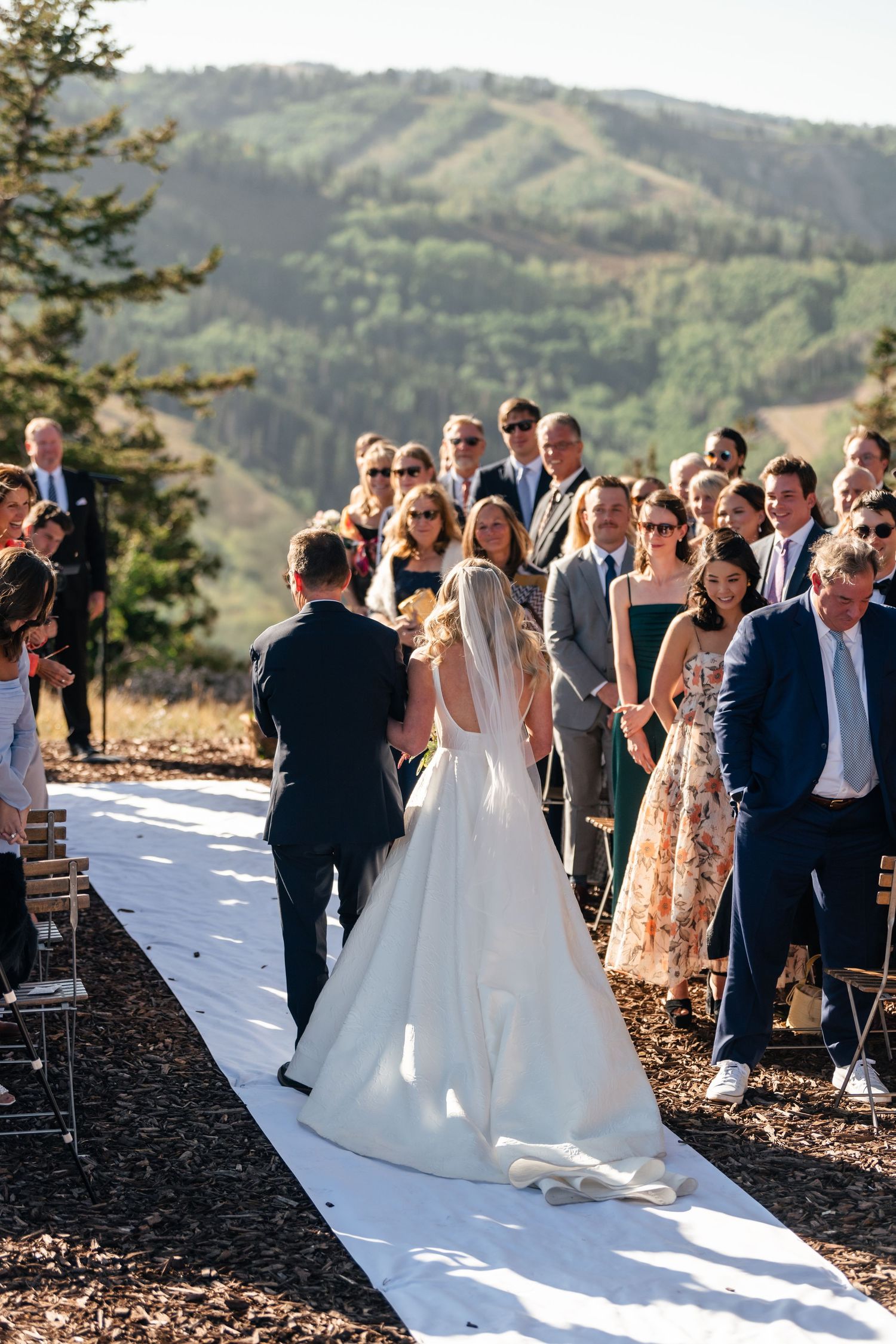 Wedding ceremony guests gather outdoors on a scenic mountain overlook as a couple walks down the white aisle runner.