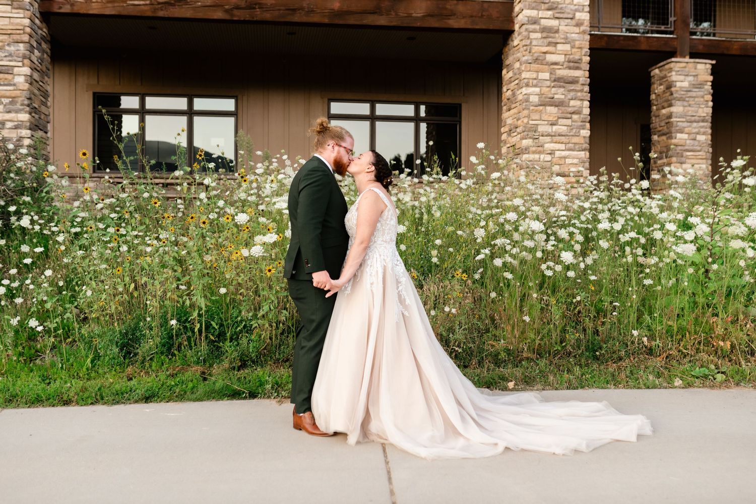 A bride in a flowing blush pink wedding gown poses outdoors against a rustic stone building with wildflowers. Wolf Creek Preferred Vendor.
