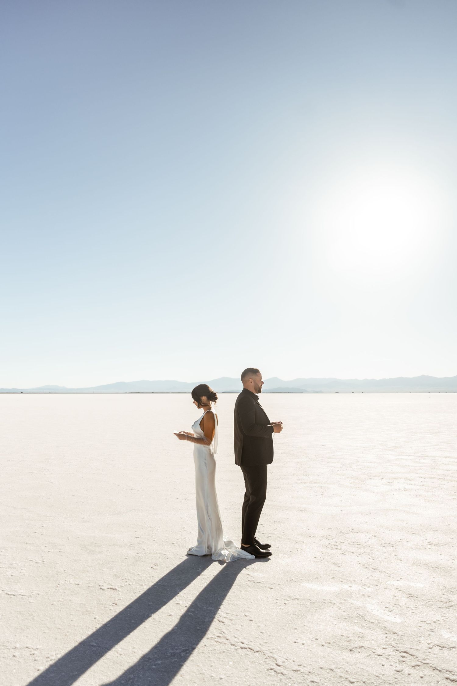 Wedding couple casting long shadows on salt flats during sunset with mountains in the distance.