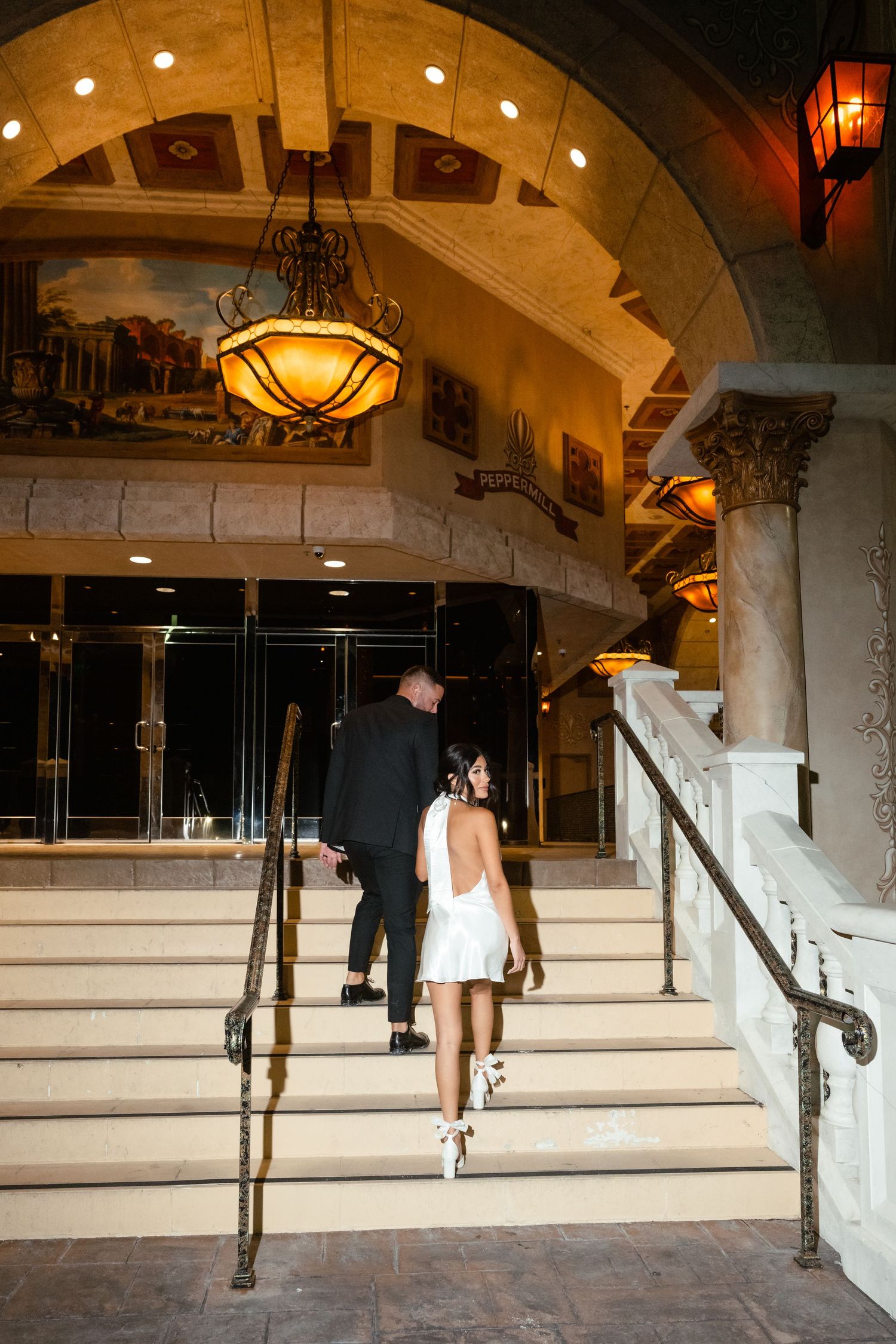 A couple ascends elegant stone stairs in an ornately decorated venue with warm lighting.