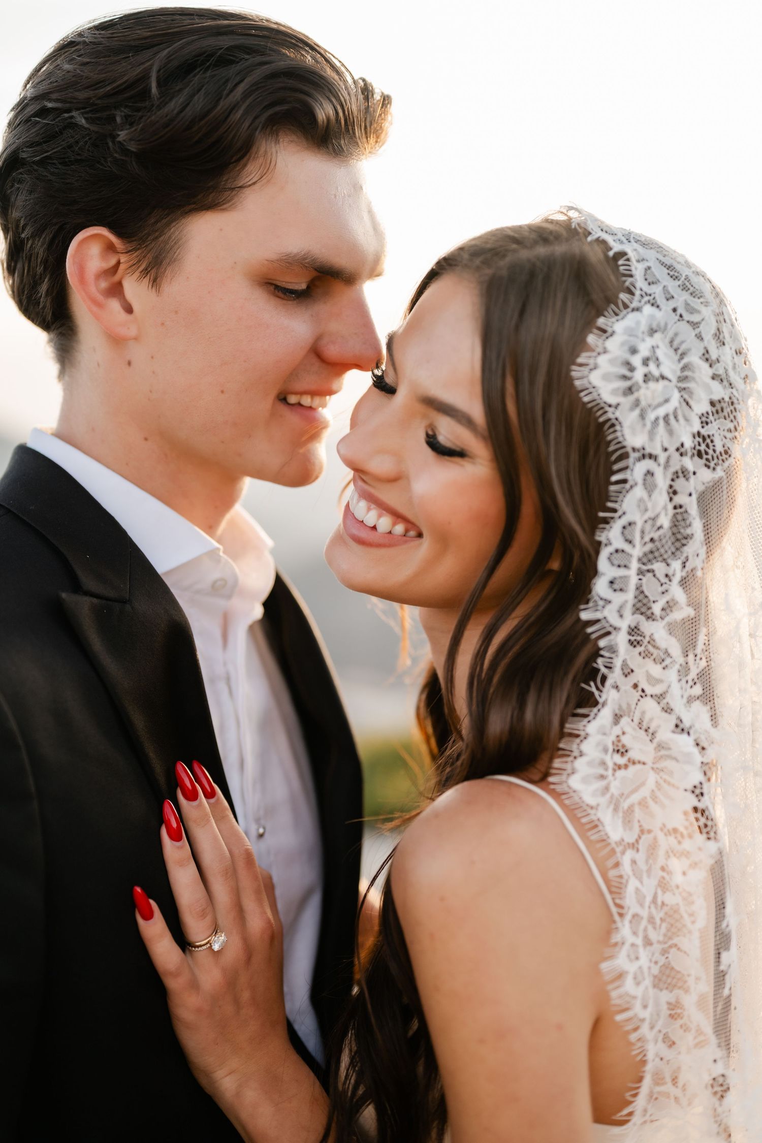 Smiling bride in lace veil and groom share a joyful forehead touch in golden sunlight.