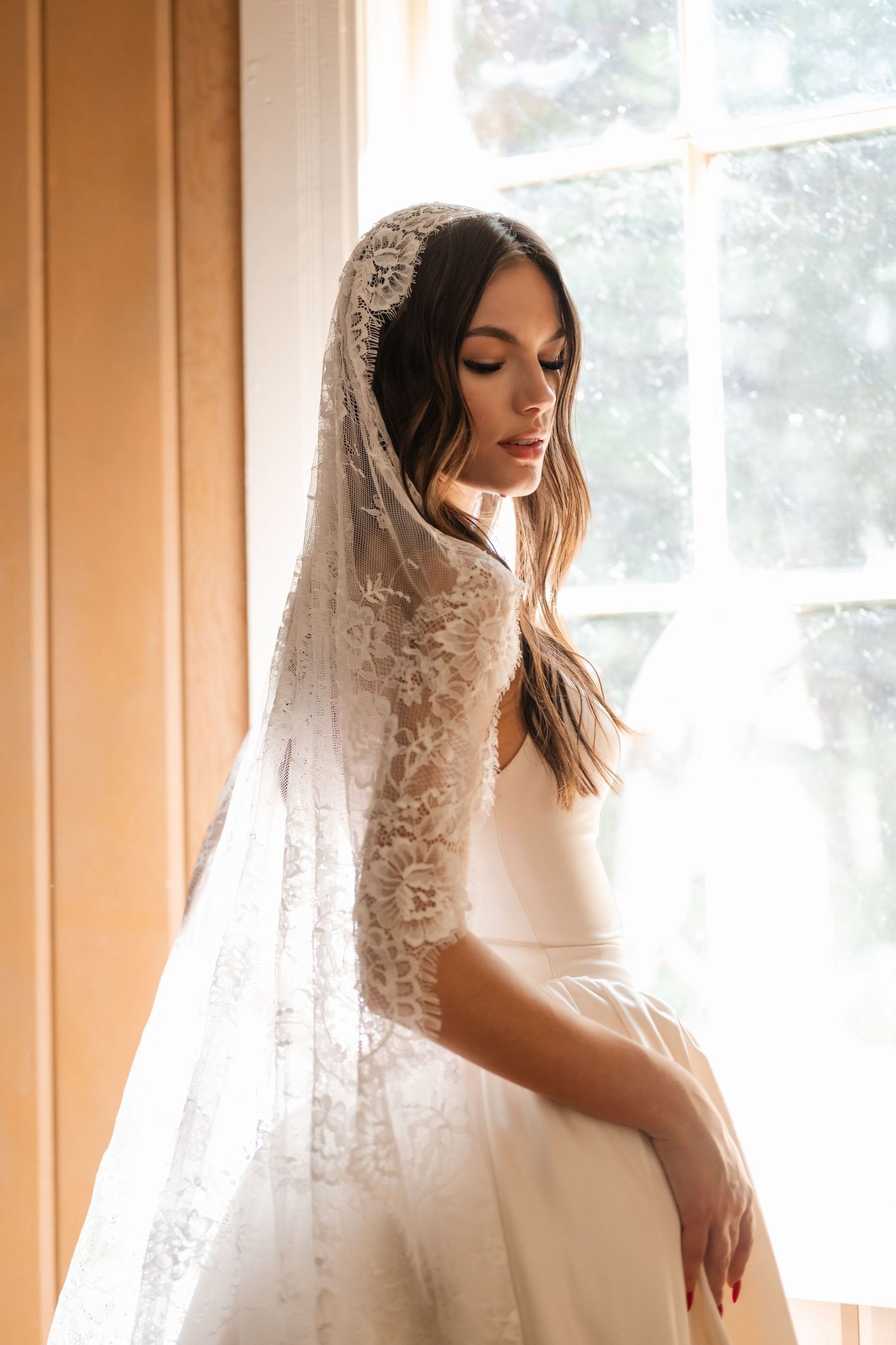 Bride wearing a white gown and intricate lace mantilla veil standing by a sunlit window.