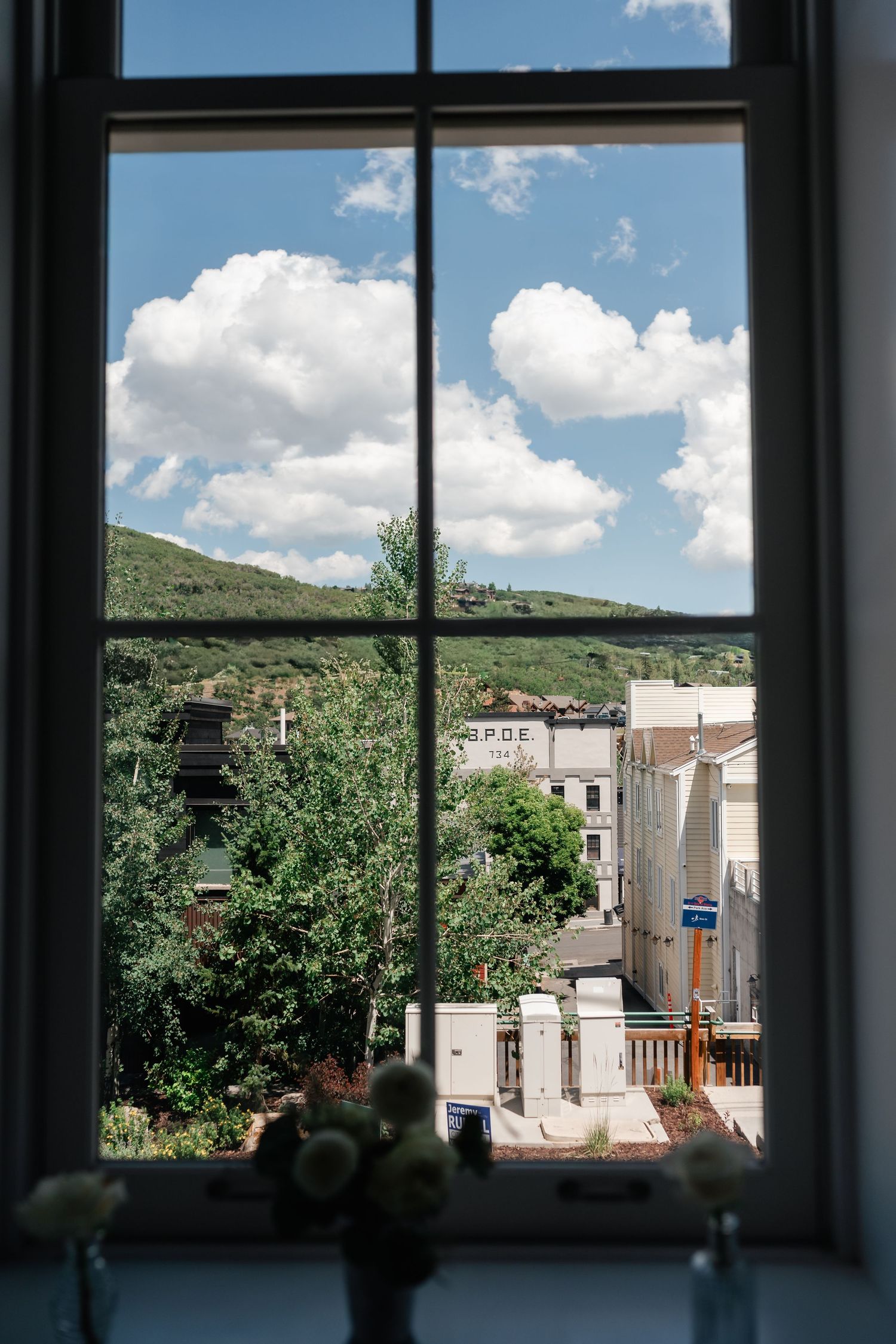 View through a window showing green hills and buildings under a blue sky with white puffy clouds.