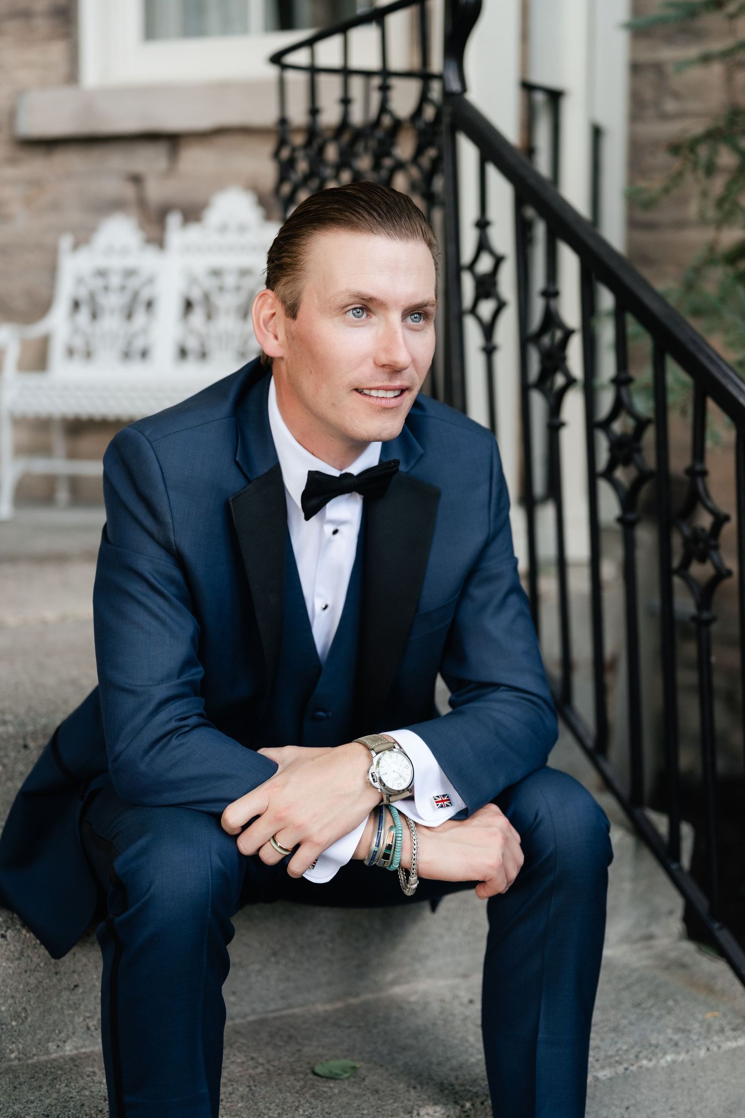 Man in navy tuxedo and bow tie sitting on stone steps with wrought iron railing.