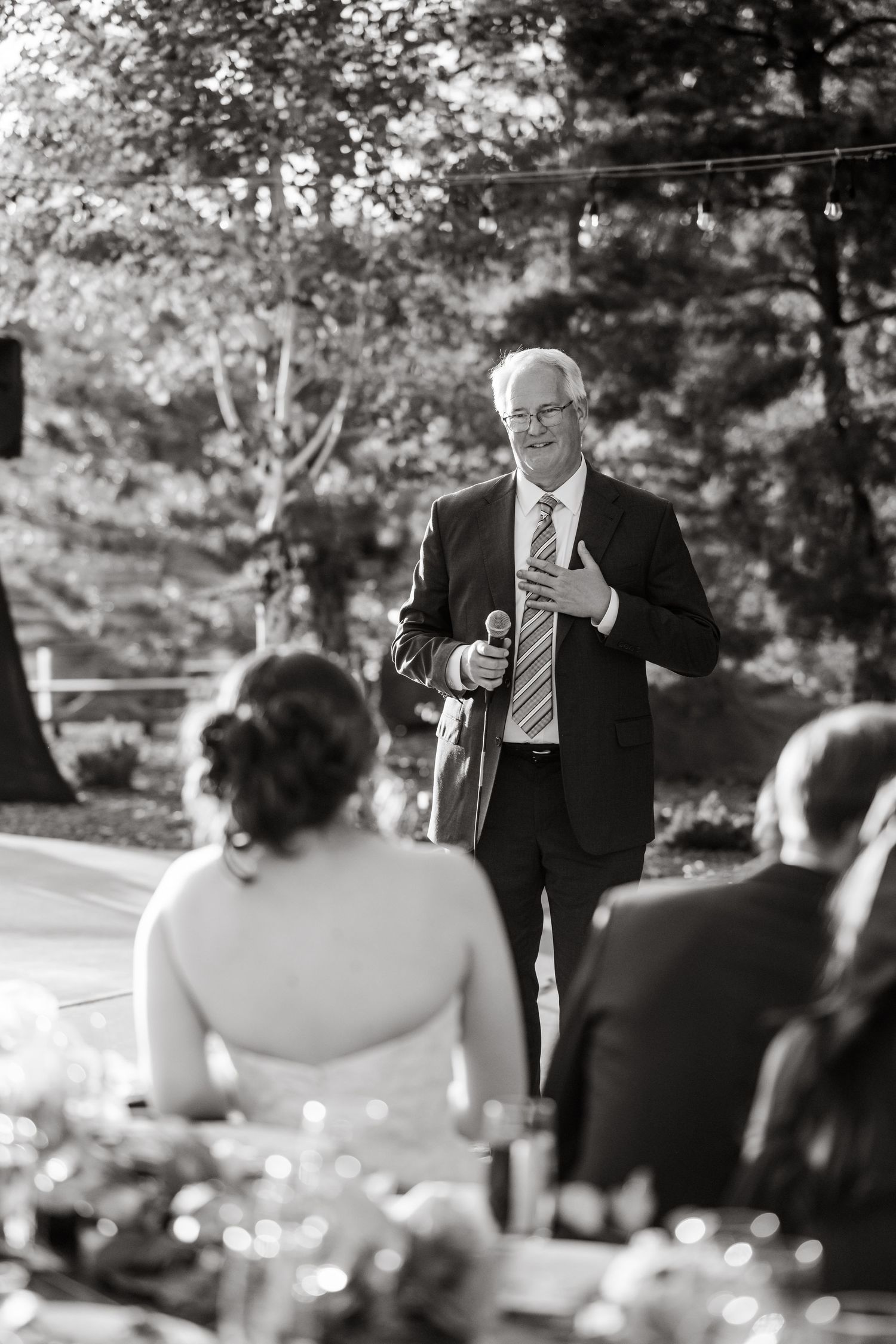 Wedding guests seated at outdoor tables listening to speeches during a reception.