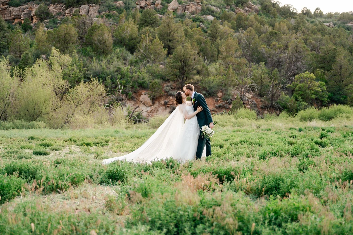 Couple in wedding attire embraces in a scenic green meadow surrounded by rolling hills and natural landscape.