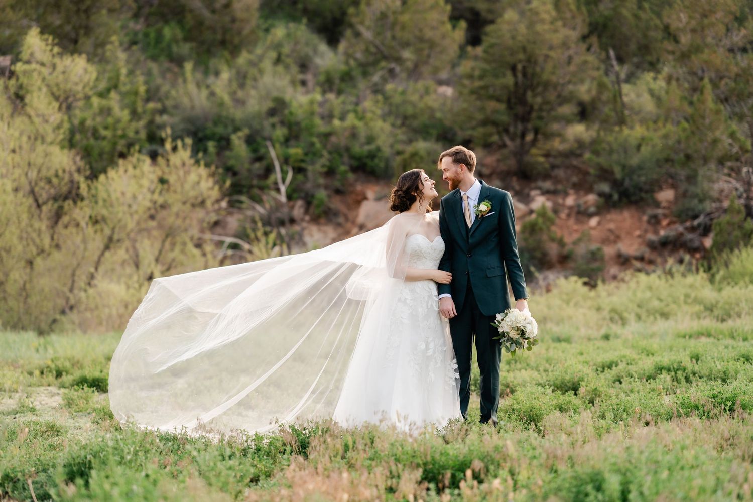 A couple stands in a green meadow as the bride's veil flows dramatically in the wind.