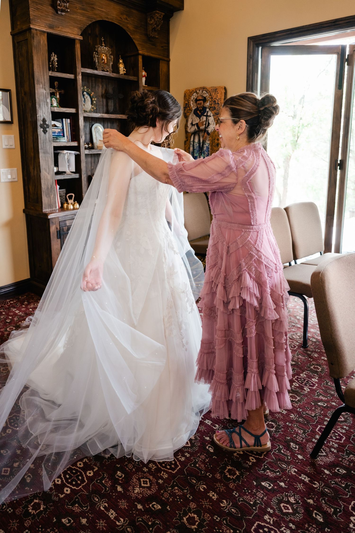 A person in a dusty pink lace dress helps adjust a flowing white wedding gown in an elegant room with wooden shelving.