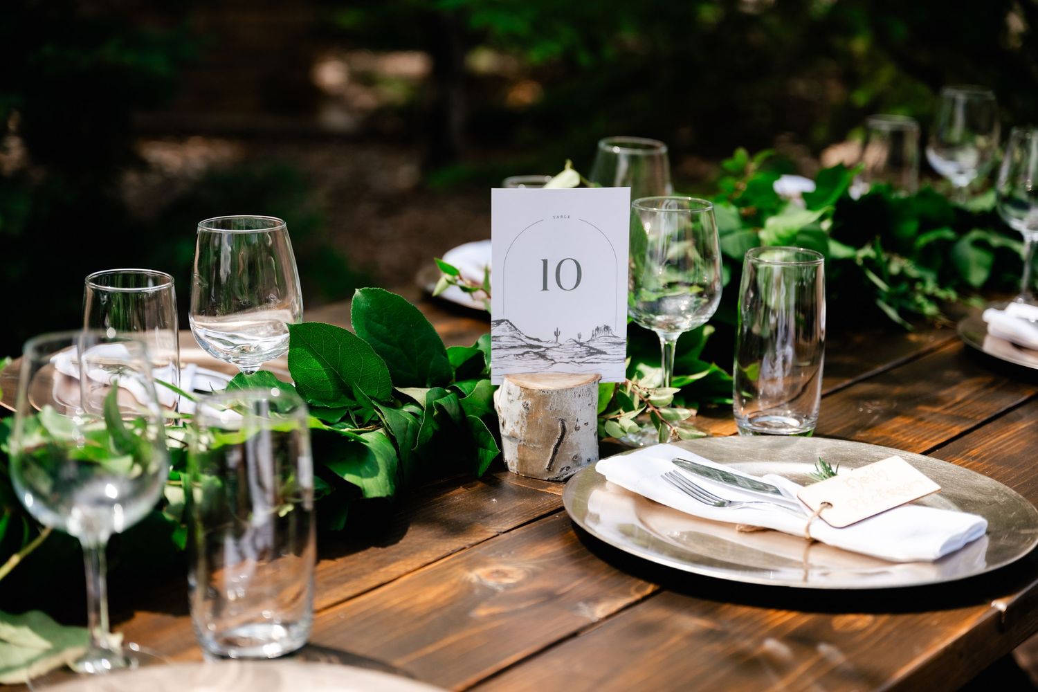 Elegant table settings with wine glasses and greenery decorations at rustic wooden reception table.