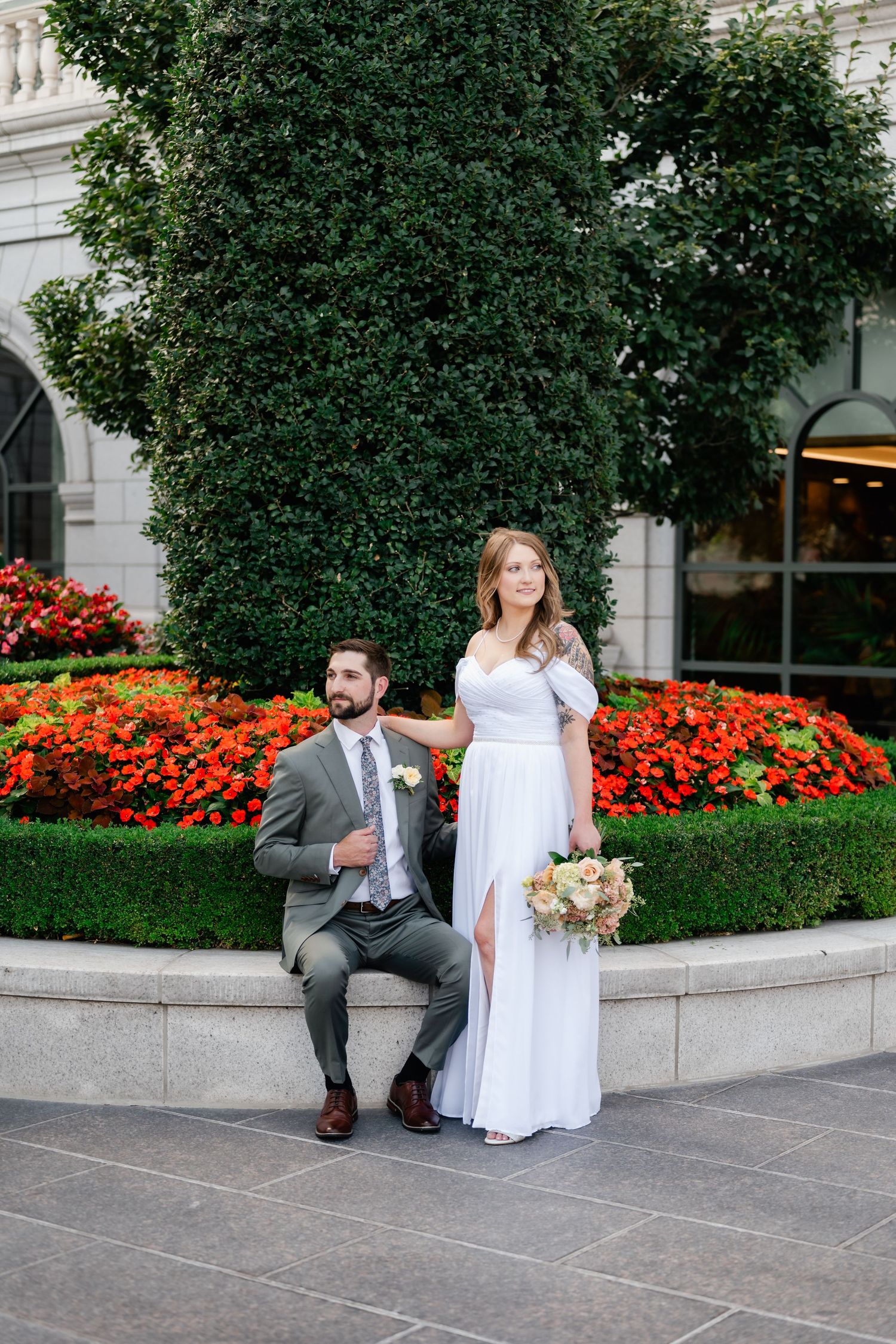 A couple poses in wedding attire in front of a garden with vibrant orange flowers and green foliage.