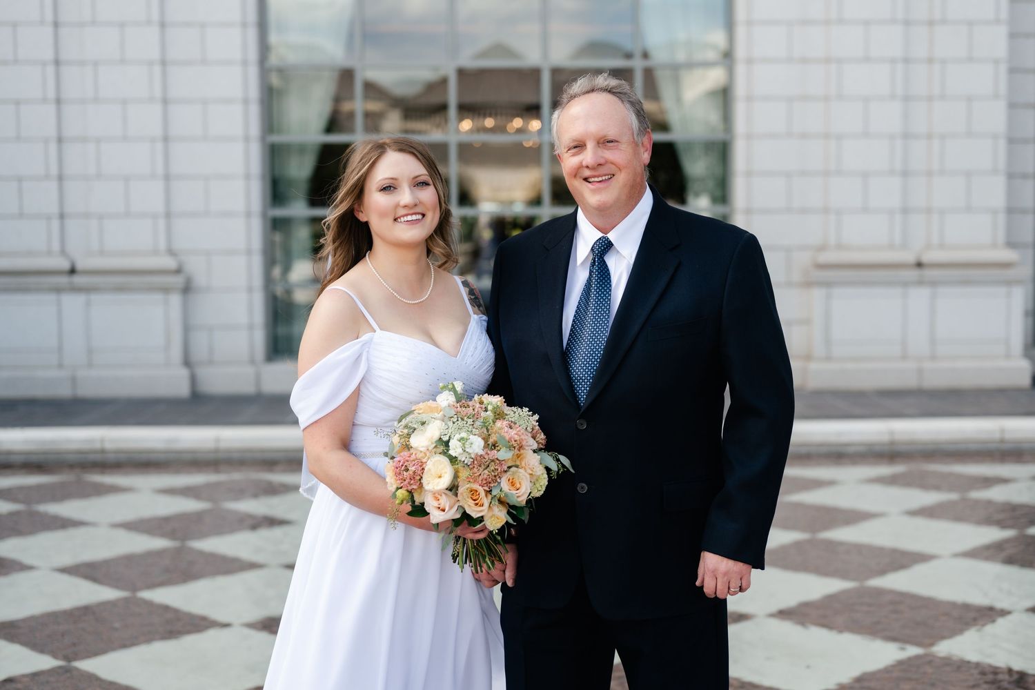 A bride in a white off-shoulder gown holding a peach and white bouquet stands with a companion on checkered marble steps.