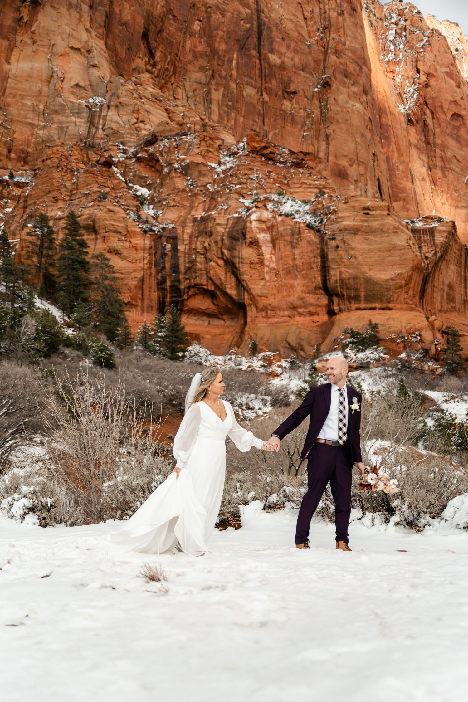 A couple in wedding attire poses in the snow against dramatic red canyon walls at sunset.