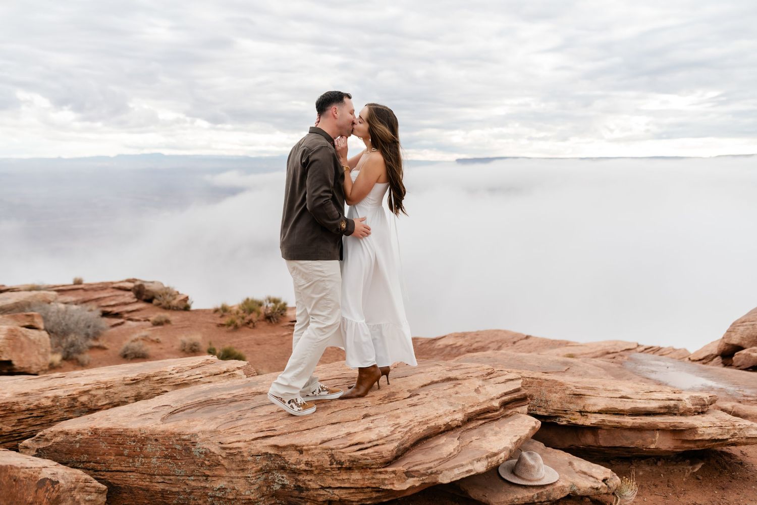 A couple shares intimate moments while standing on red rock formations under an overcast sky.