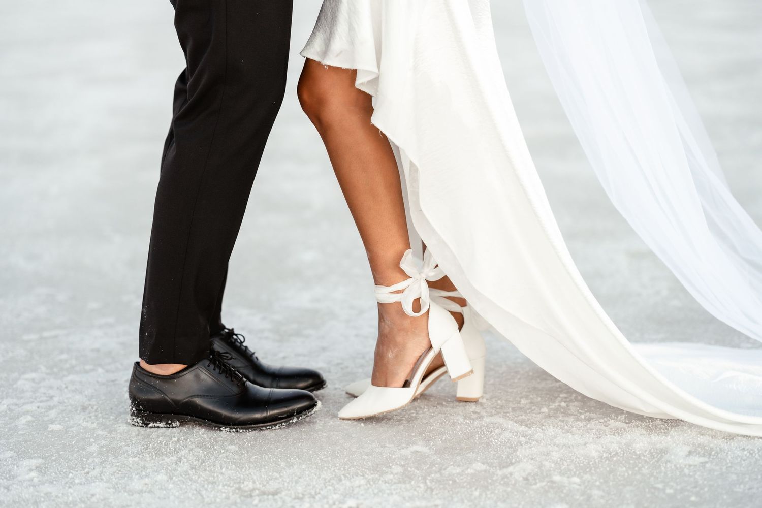 Close-up of formal wedding shoes and dress hem on white salt flat surface.