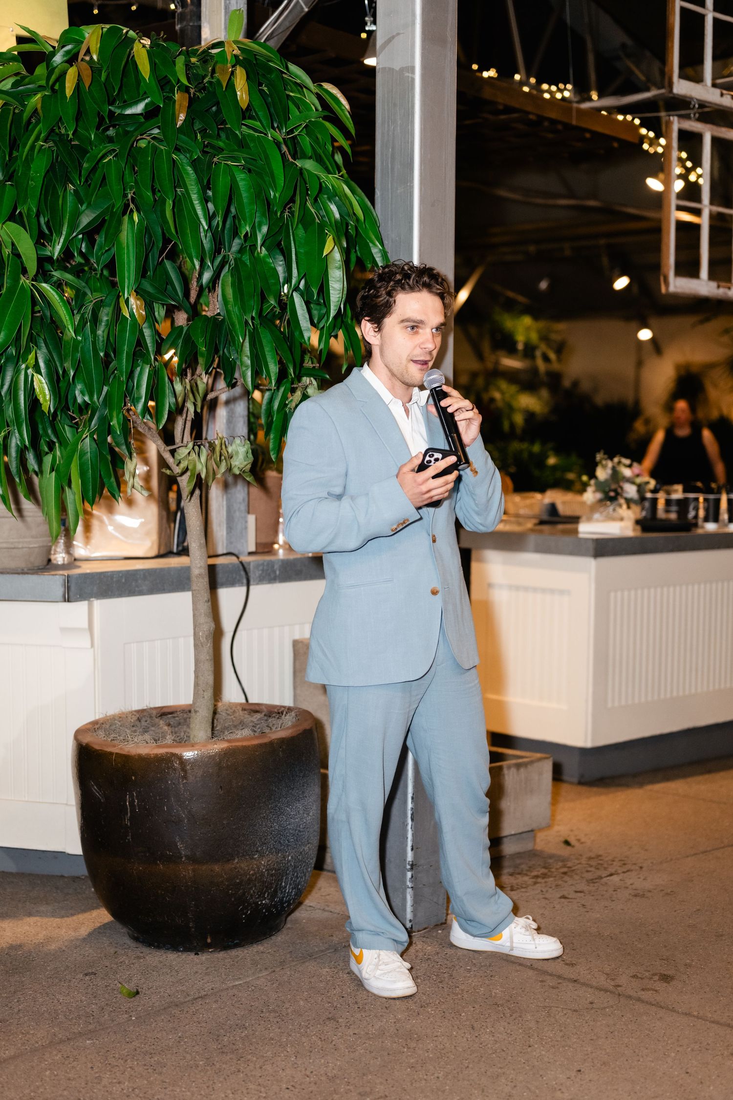 A figure in a light blue suit stands near a potted plant at an indoor event venue.