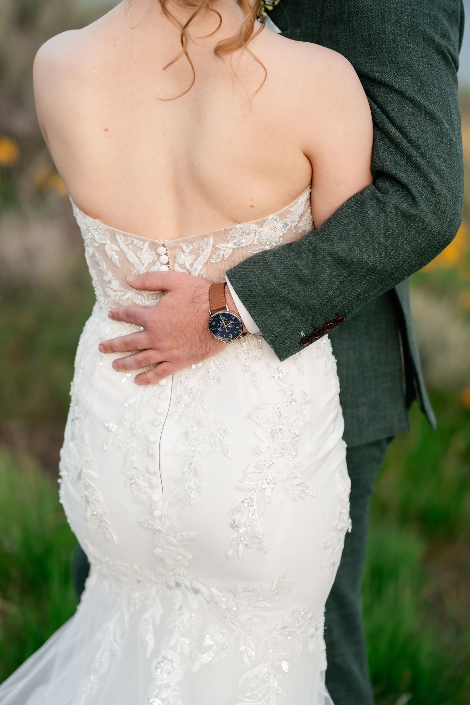 Close-up detail of bride's lace dress and groom's hand with watch.