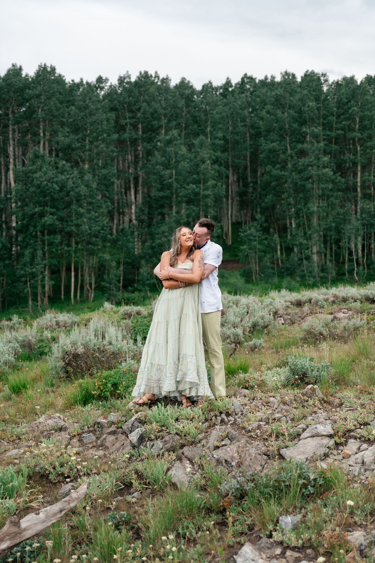 Two people embrace on a rocky hillside with a dense pine forest backdrop in a romantic mountain setting. Utah mountain engagement session