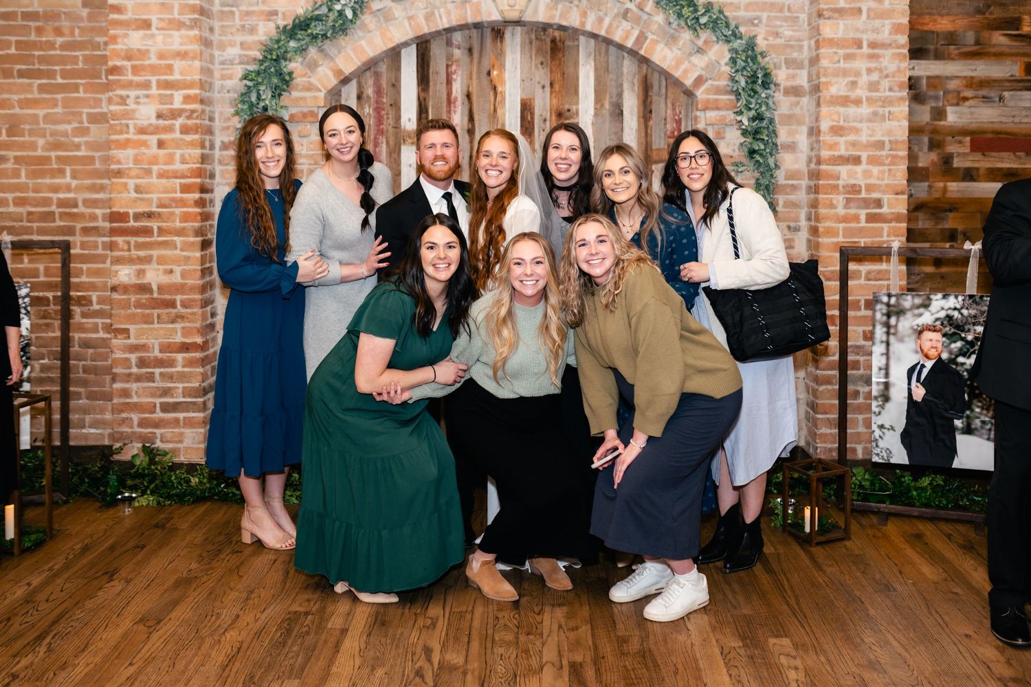 A group poses together against a rustic brick wall decorated with greenery garland in an indoor venue.