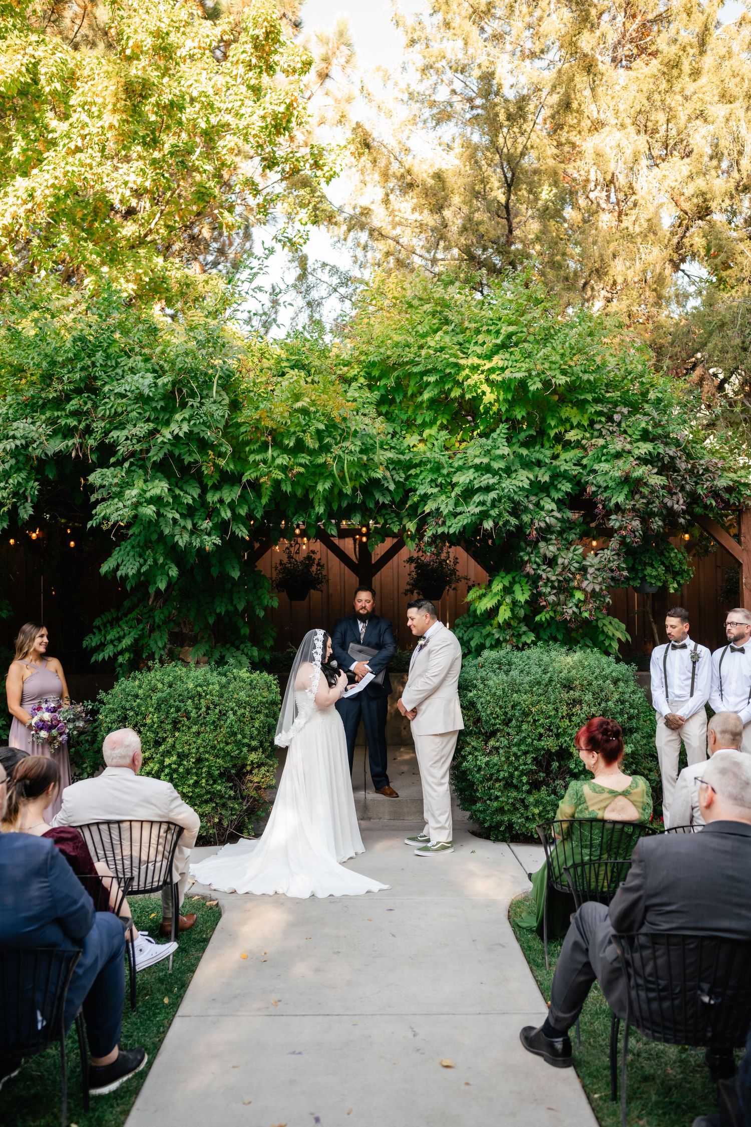 An outdoor garden wedding ceremony takes place on a paved path surrounded by lush greenery and guests seated on chairs.