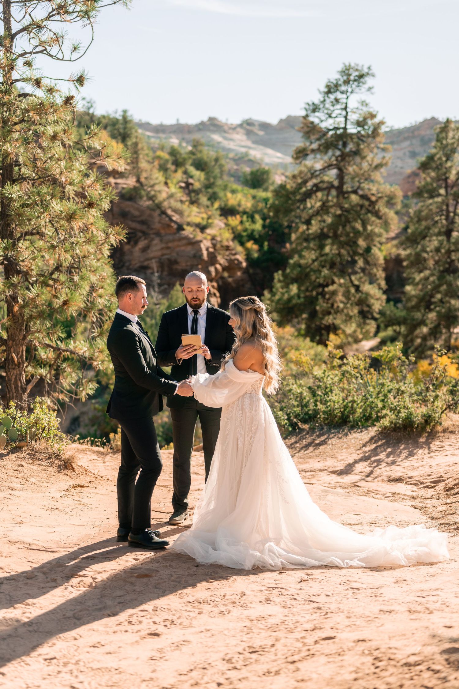 A wedding ceremony unfolds on a scenic mountain trail surrounded by pine trees and desert landscape.