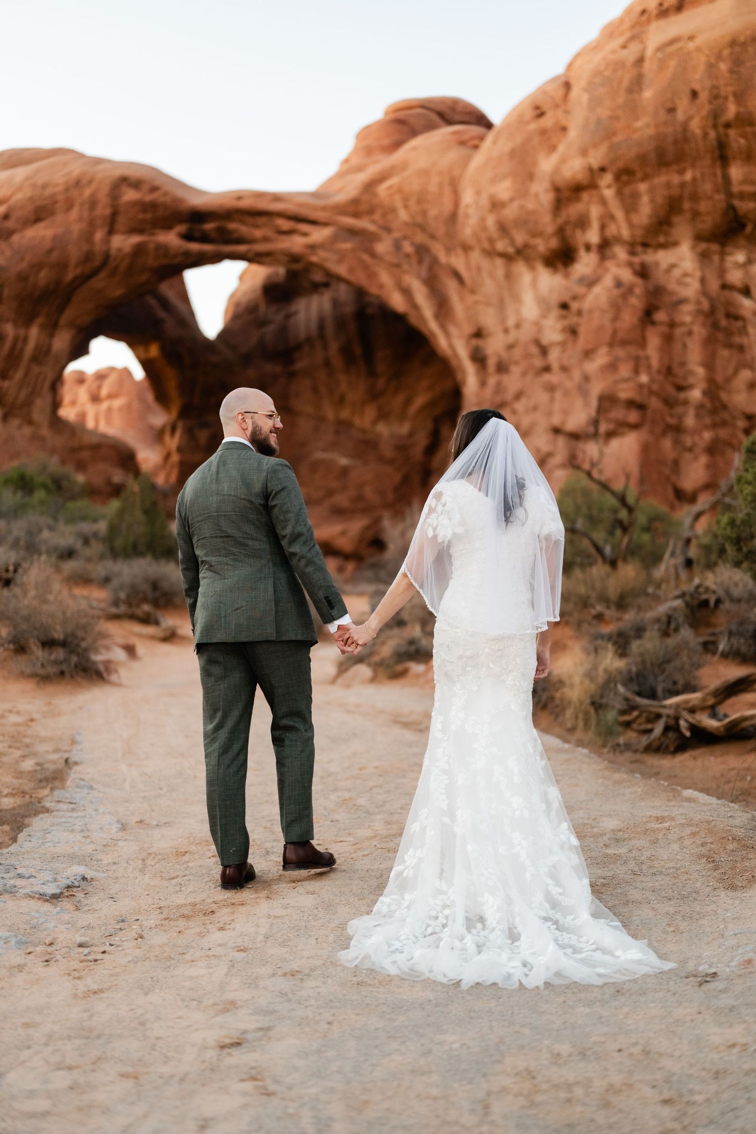 Wedding couple walking along desert trail with majestic red rock arches in background.