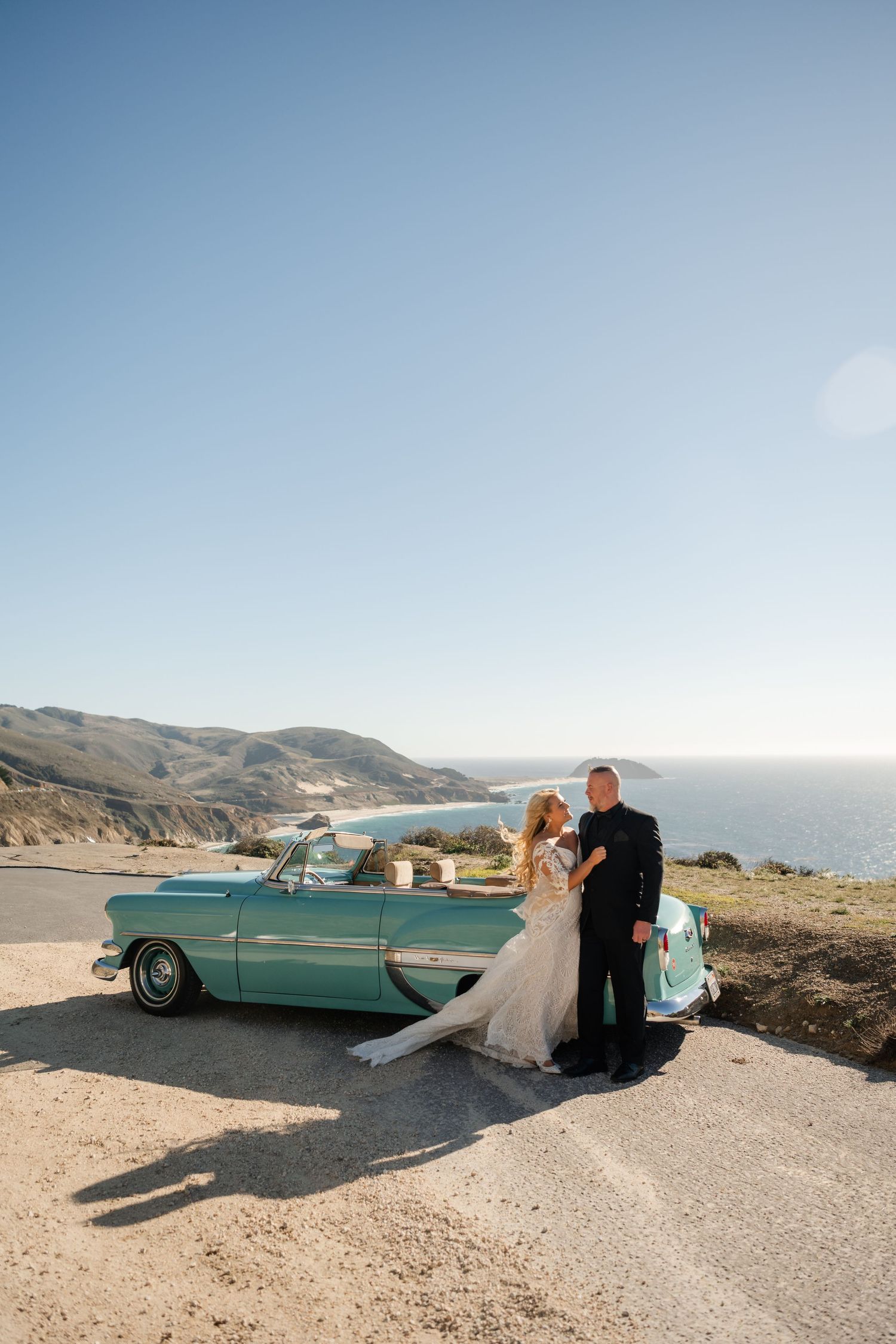A vintage turquoise car parked on a coastal cliff overlooking the ocean during a wedding photoshoot.
