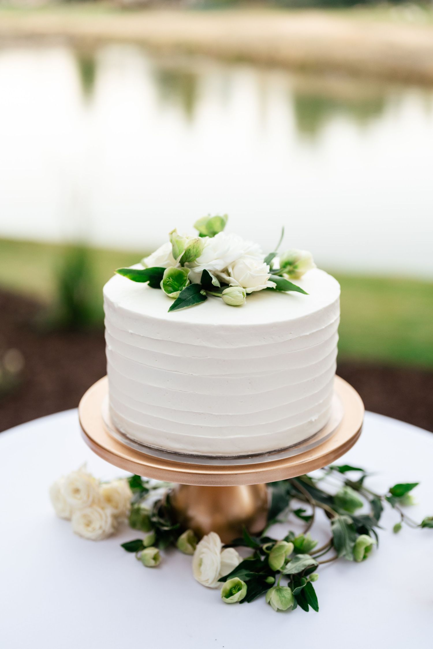 An elegant white wedding cake decorated with fresh greenery sits on a gold cake stand.