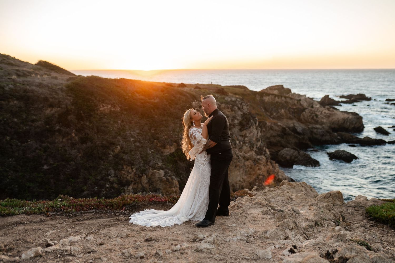 Couple embracing on rocky coastal cliffs at sunset with ocean waves crashing below.