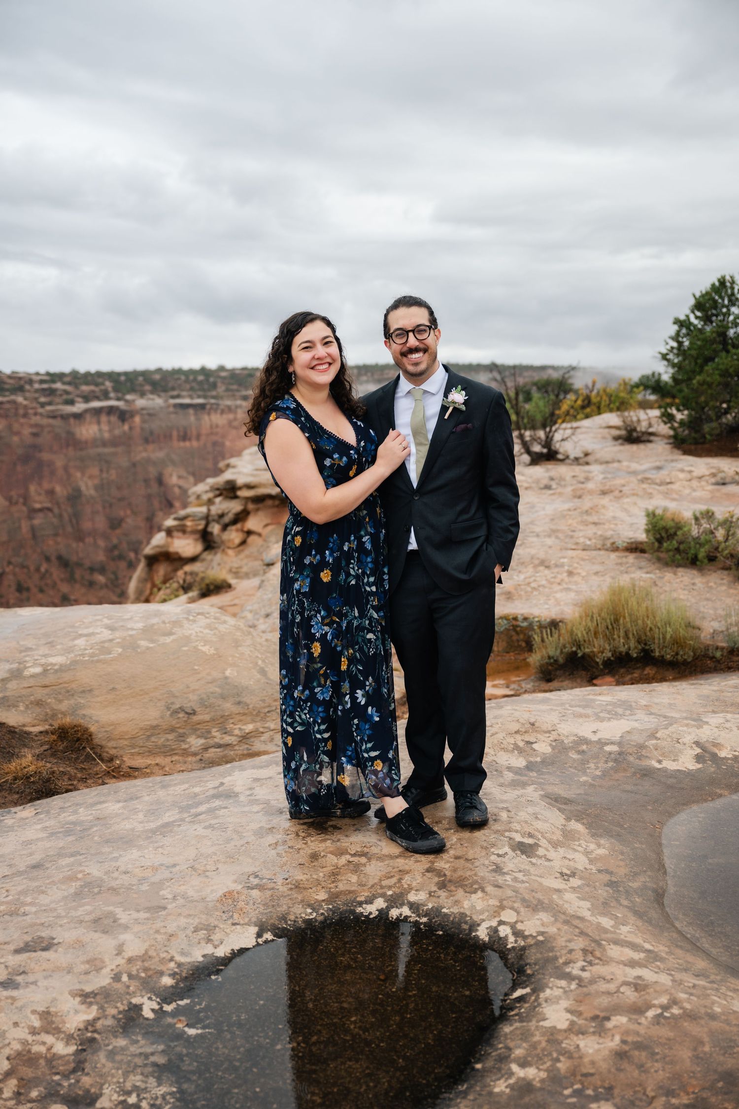 Wedding couple poses on rocky terrain overlooking a vast desert canyon under cloudy skies.