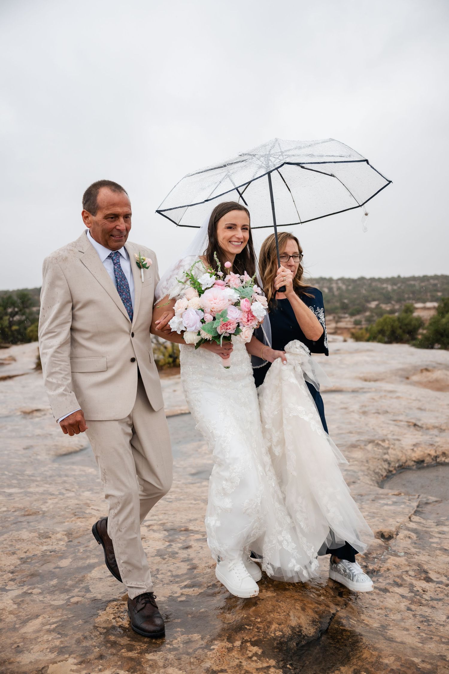 Wedding party walking on rocky terrain with clear umbrellas and pink bouquet.