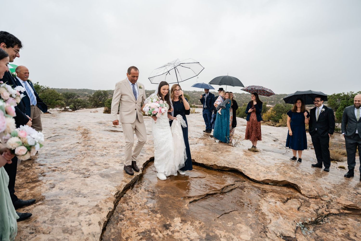 Wedding guests gather on a rugged desert cliff with umbrellas during an overcast day.