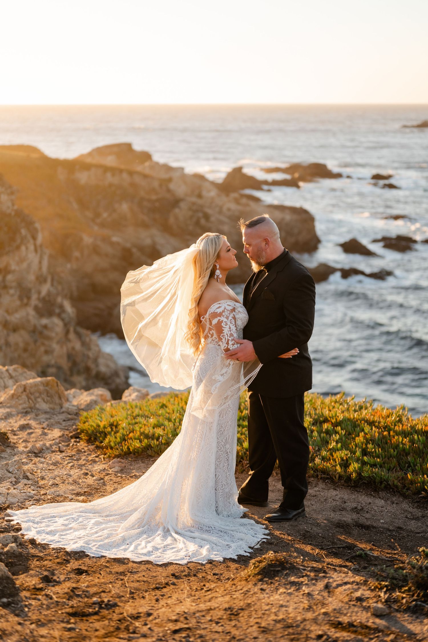 Newlyweds share an intimate moment on a scenic coastal cliff at golden hour.