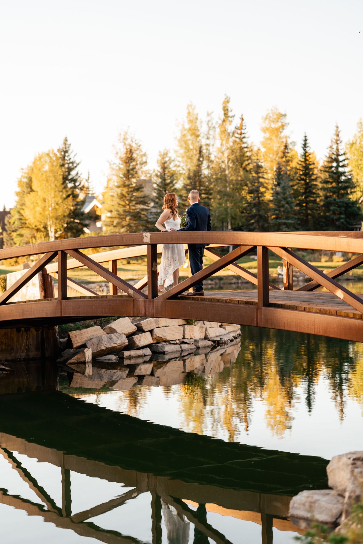 A wooden bridge spans across a serene pond with evergreen trees reflecting in the water.
