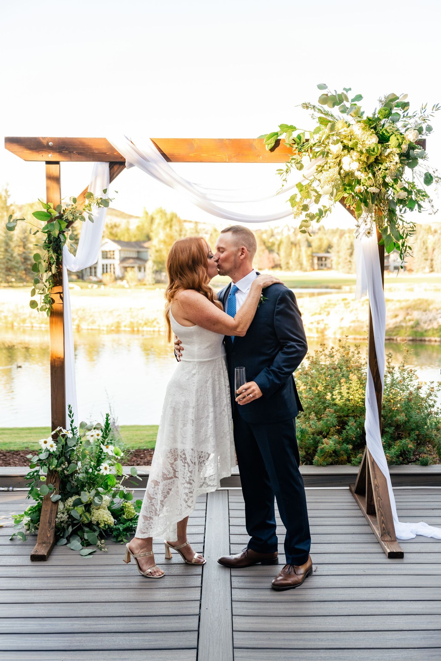 A couple in wedding attire shares a romantic moment on a wooden deck overlooking a lake at sunset.